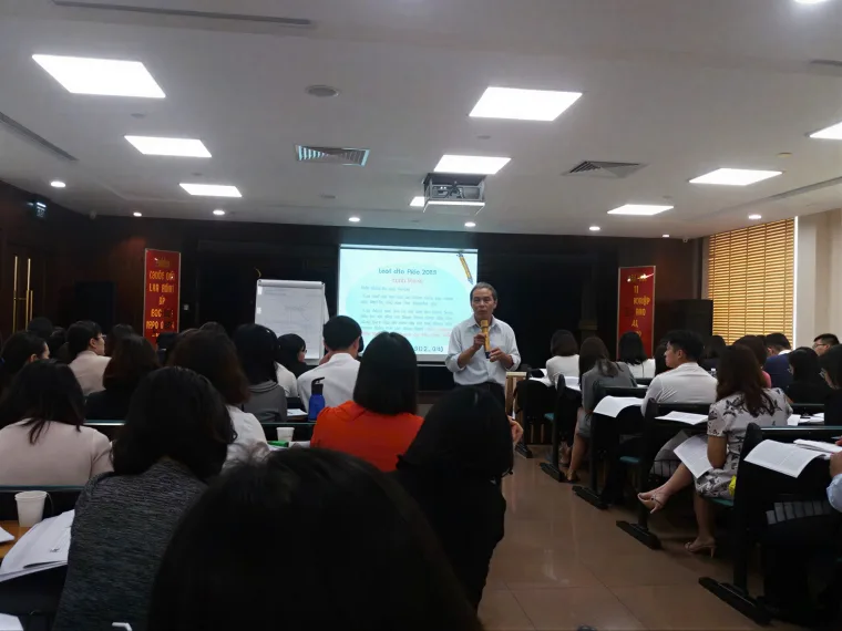 Giảng viên cầm micro giảng bài trước lớp, học viên ngồi theo dõi và ghi chép trong khóa đào tạo nghiệp vụ.